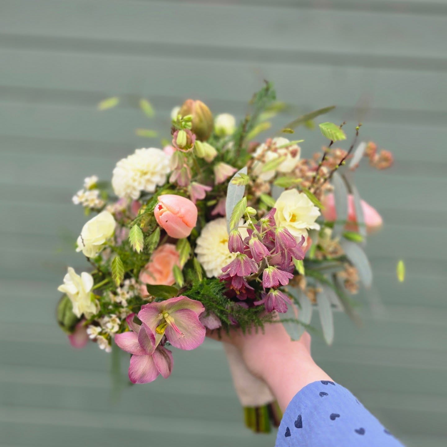 Buket med rosa tulipaner, lilla klokkeformede blomster og lyse blomster foran bodens grønne væg