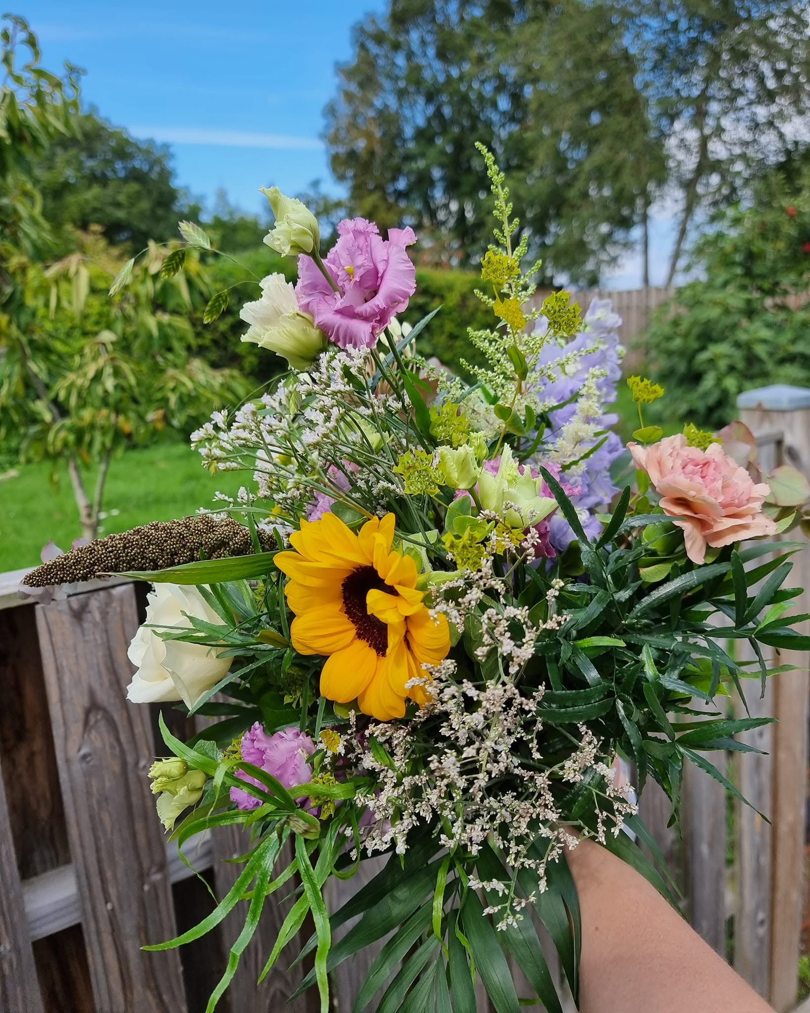 Buket med en gul solsikke, lilla og lyse blomster samt grønt i haven